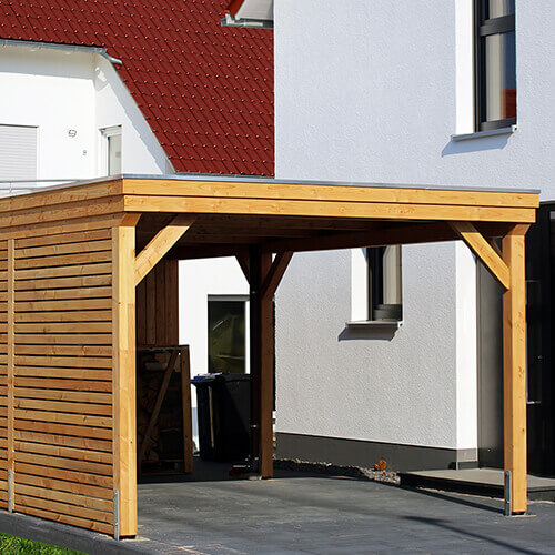 Ein moderner Carport aus Holz, der sicher an der Seite eines weißen Hauses befestigt ist. Der Carport hat ein flaches Dach und bietet Schutz für Fahrzeuge. Die Holzstruktur ist stabil gebaut und gut verankert.
