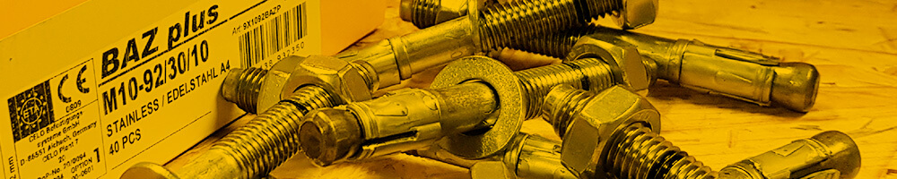 Eine Nahaufnahme von mehreren Bolzenankern, die auf einer Oberfläche liegen. Im Hintergrund ist eine gelbe Verpackungsschachtel mit der Aufschrift "BAZ plus M10-92/30/10" zu sehen. Die Bolzenanker sind aus Edelstahl und bereit zur Verwendung.