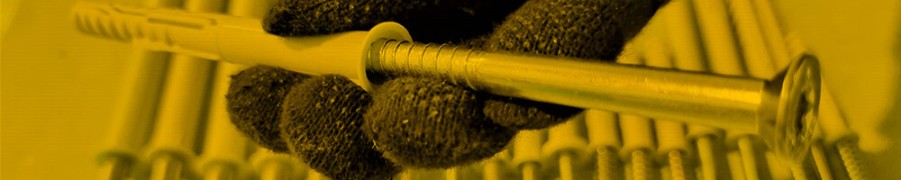 Eine behandschuhte Hand hält einen langen Rahmendübel, auch Fassadendübel genannt, mit mehreren weiteren Dübeln unscharf im Hintergrund. Das Bild ist in einem gelblichen Farbton gehalten und zeigt die Detailansicht des Befestigungselements.