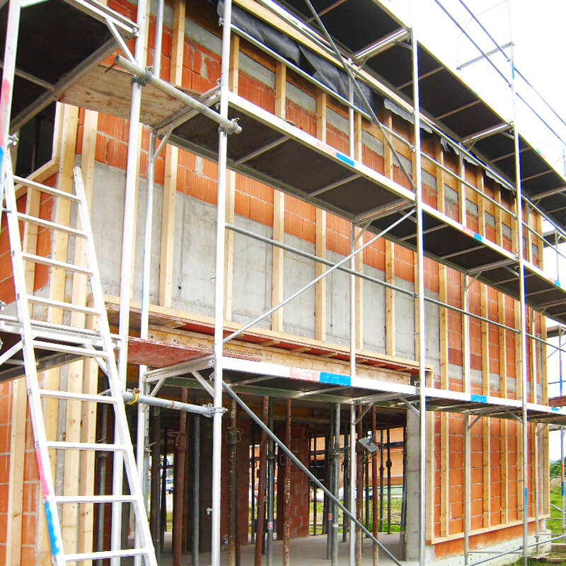 Eine Baustelle mit einem im Bau befindlichen Gebäude, das von einem Baugerüst umgeben ist. Das Gerüst ist sicher an der Fassade des Gebäudes befestigt. Die Fassade zeigt sichtbare Holzlattungen, die zur Befestigung der Dämmung oder Verkleidung verwendet werden.