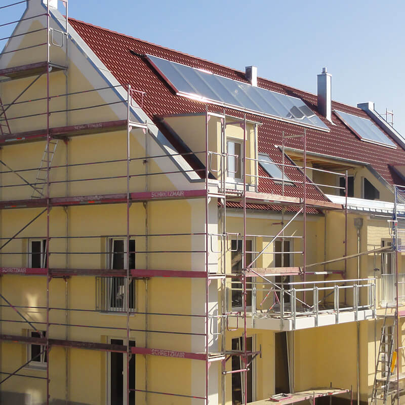 Ein mehrstöckiges Wohnhaus mit gelber Fassade und rotem Dach, das von einem Baugerüst umgeben ist. Das Gerüst ist sicher an der Fassade befestigt, um Arbeiten an der Außenwand und dem Dach zu ermöglichen.