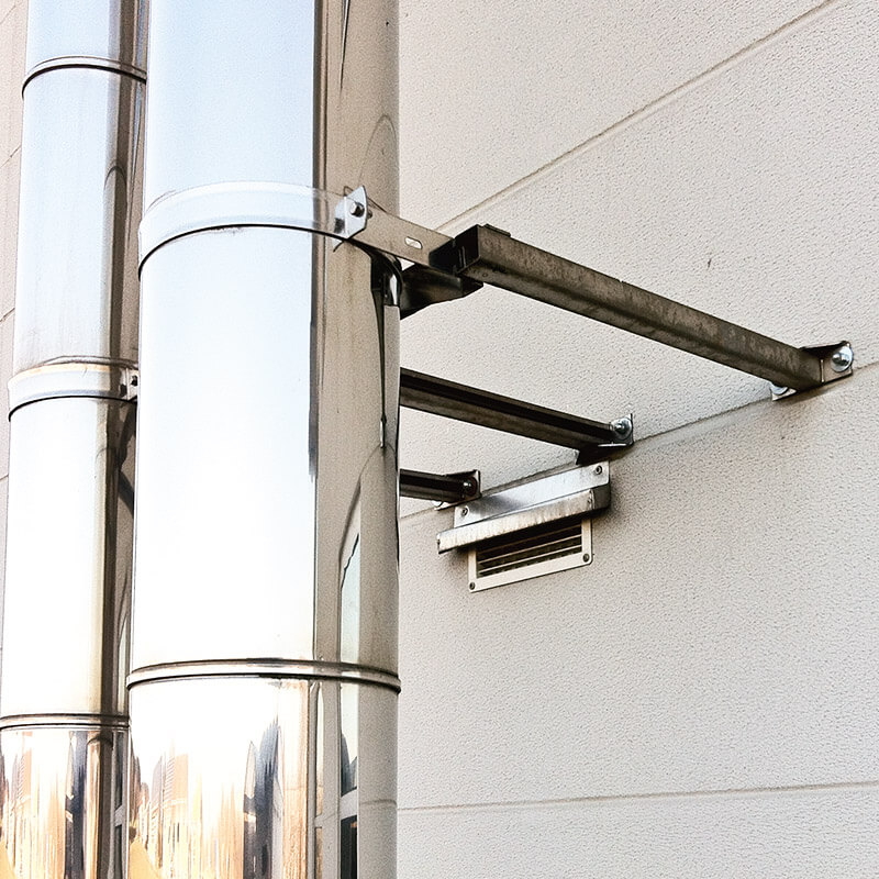 Ein großes, glänzendes Metallrohr, das mit robusten Halterungen an einer Außenwand befestigt ist. Die Halterungen sind sicher an der Wand montiert und stützen das Rohr, das Teil einer industriellen Belüftungs- oder Abgasanlage ist.