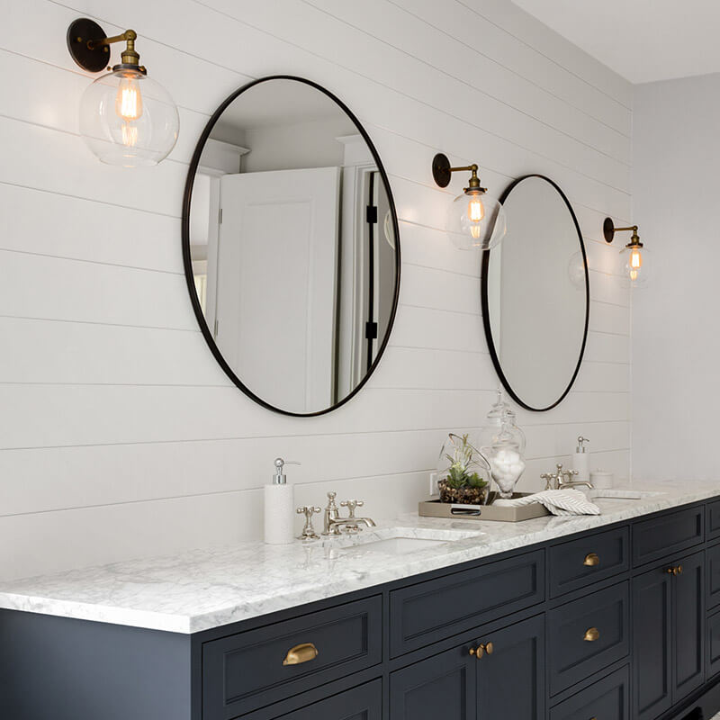 Ein modernes Badezimmer mit einer langen Waschtischplatte aus Marmor, die sicher an der Wand befestigt ist. Über der Waschtischplatte sind zwei runde Spiegel an der Wand montiert, flankiert von stilvollen Wandleuchten mit sichtbaren Glühbirnen