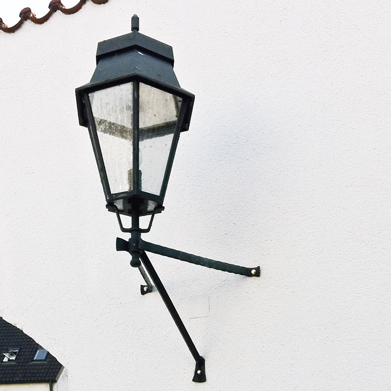 Eine antike Außenlaterne, die mit robusten Metallhalterungen an einer weißen Hauswand befestigt ist. Die Halterungen sind sicher an der Wand verschraubt, um die Laterne zu stützen und zu sichern.