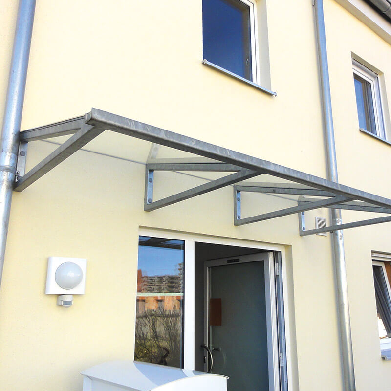 Ein Vordach aus Metall und Glas, das über dem Eingang eines Hauses angebracht ist. Die Metallkonstruktion des Vordachs ist sicher mit chemischen Mörteln an der Hauswand verankert, um Stabilität und Haltbarkeit zu gewährleisten.