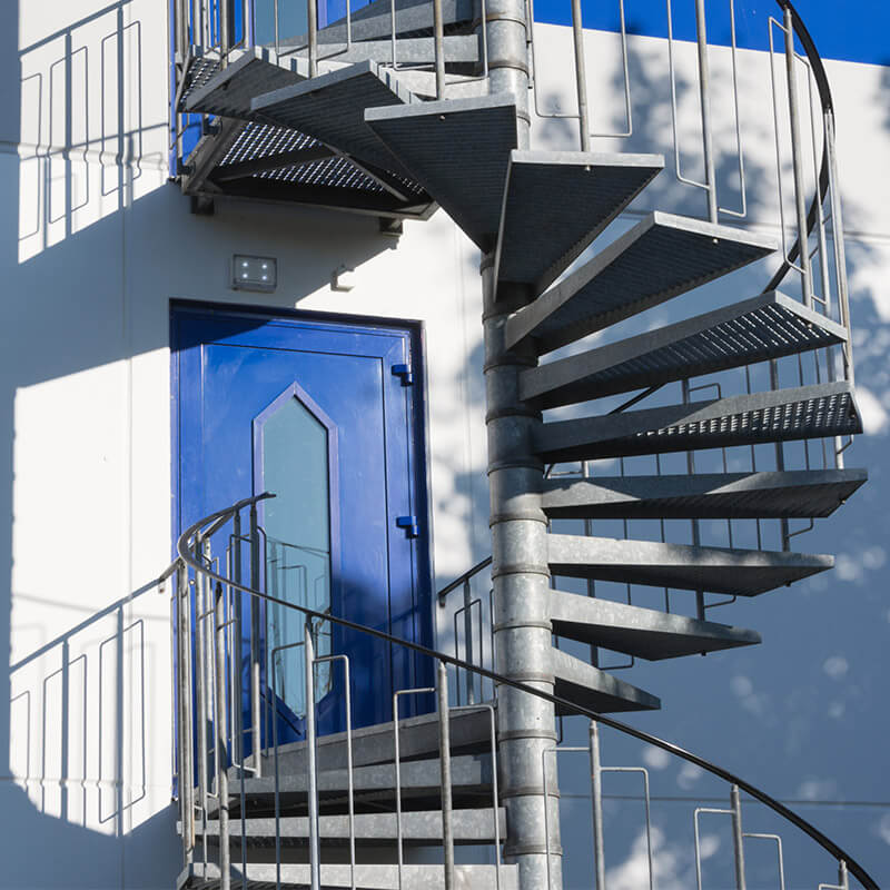 Eine metallene Wendeltreppe, die an der Außenseite eines Gebäudes befestigt ist. Die Treppe ist mit chemischen Mörteln sicher an der Wand und am Boden verankert, um eine stabile und langlebige Konstruktion zu gewährleisten.