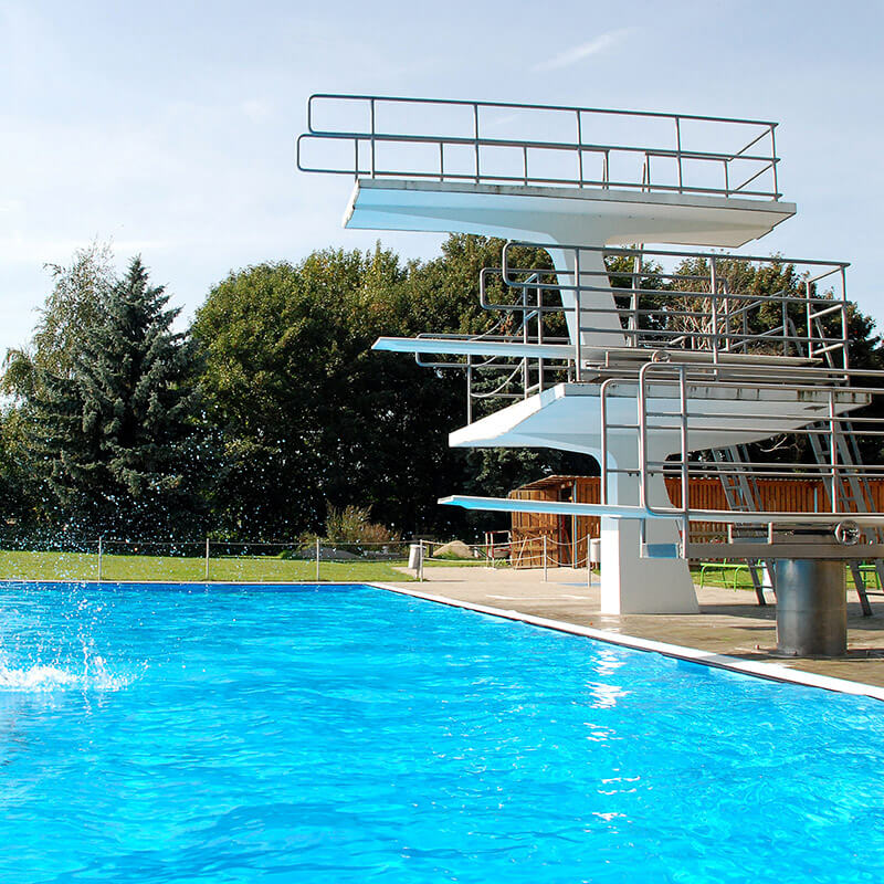 Ein Freiluftschwimmbad mit einem hohen Sprungturm, der mehrere Plattformen und Sprungbretter hat. Der Sprungturm ist sicher mit chemischen Mörteln an der Basis verankert, um eine stabile und dauerhafte Befestigung zu gewährleisten auch in Umgebungen mit hohen Belastungen wie z.B. durch Chlor oder salzige Meeresluft.