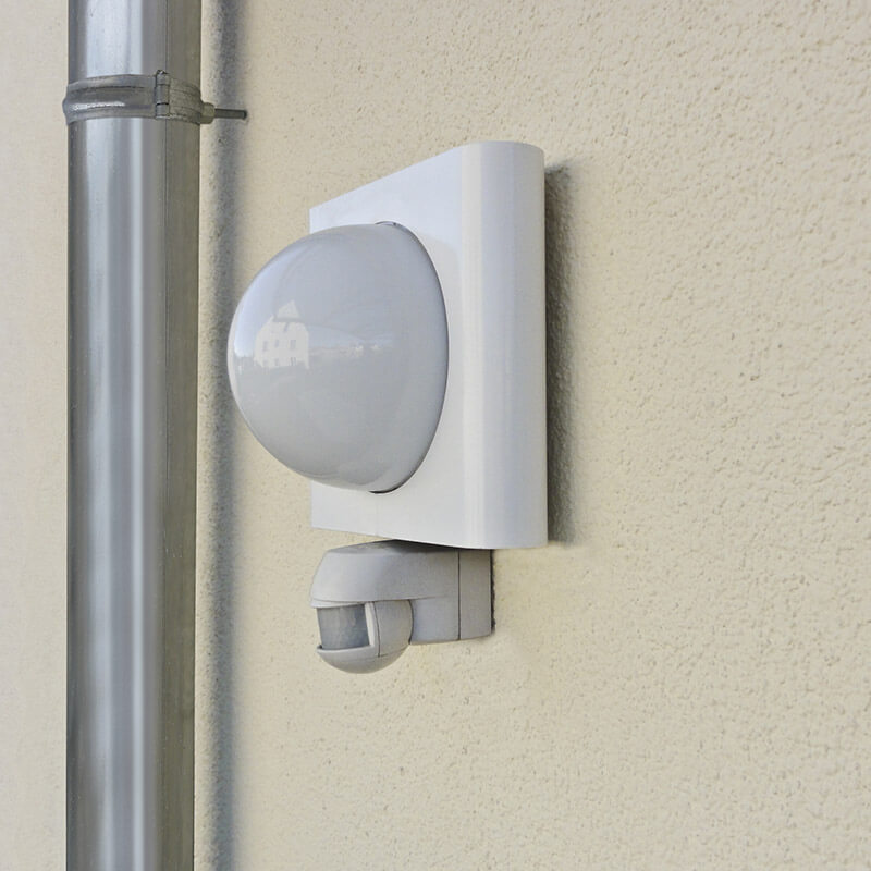 Eine Außenwand eines Gebäudes mit einem angebrachten Bewegungsmelder und einer Außenleuchte. Beide Geräte sind mit CELO-Dämmstoffdübeln an der gedämmten Fassade befestigt, um eine stabile und sichere Montage zu gewährleisten. Neben der Leuchte verläuft ein Regenfallrohr, das ebenfalls mit geeigneten Befestigungselementen fixiert ist.