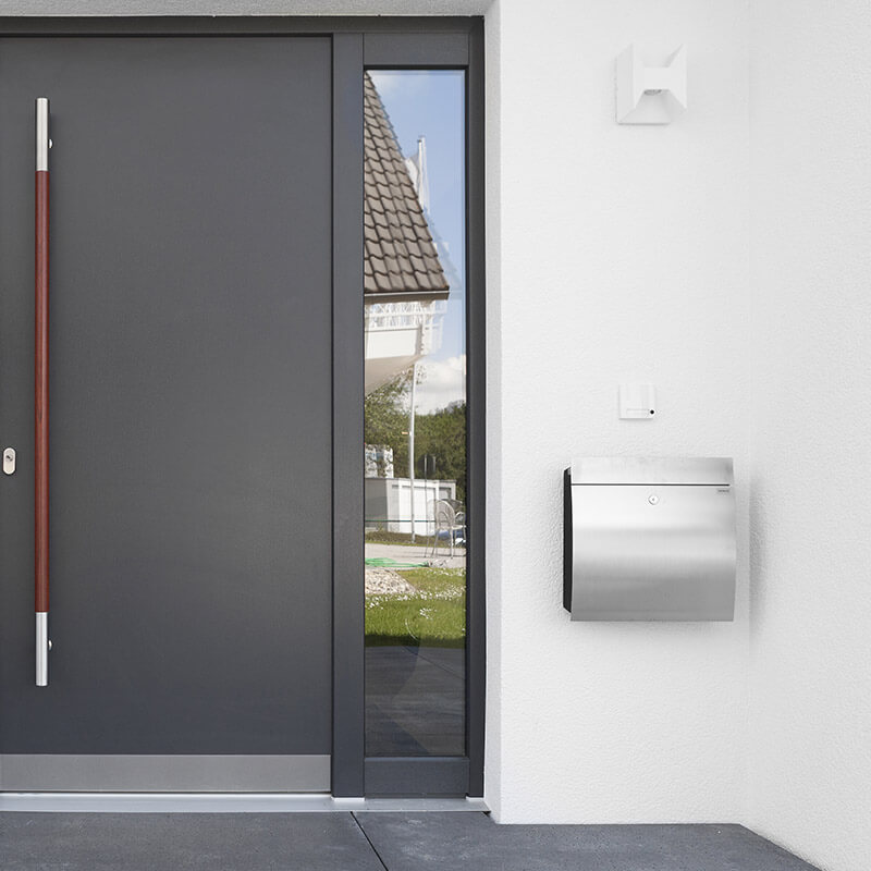 Eine moderne Hauseingangstür mit seitlichem Glaselement und einem daneben an der gedämmten Fassade befestigten Briefkasten aus Edelstahl. Der Briefkasten ist mit CELO-Dämmstoffdübeln sicher montiert, um eine stabile Befestigung an der gedämmten Hauswand zu gewährleisten.