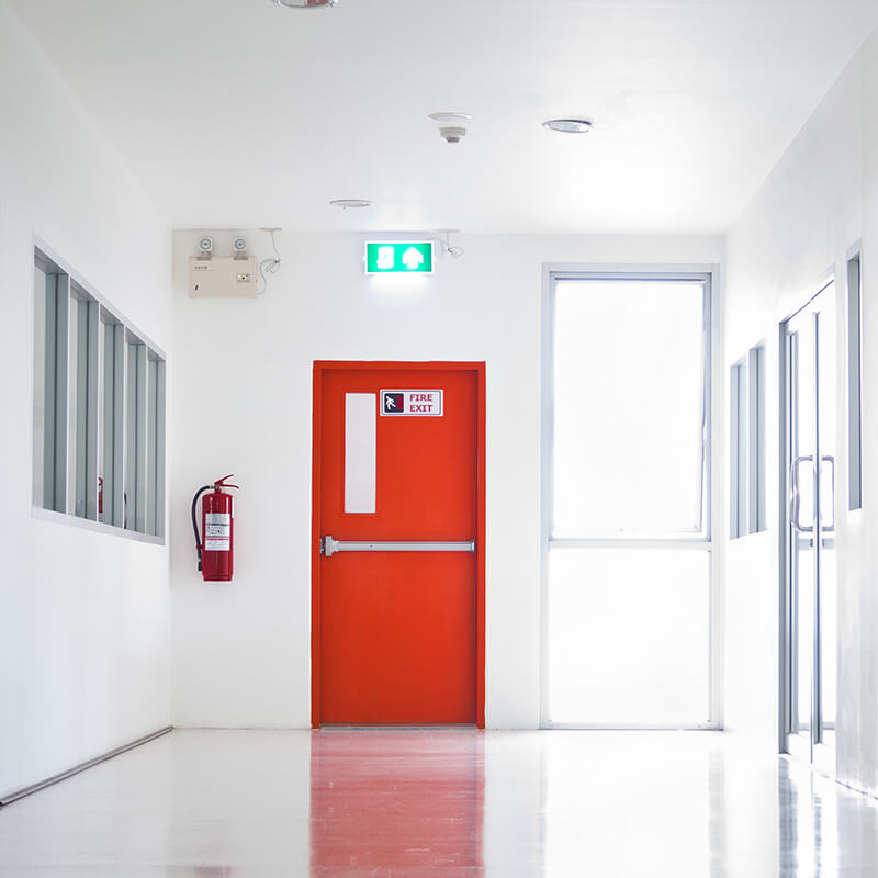 Ein heller Korridor in einem Gebäude mit einer auffälligen roten Feuertür, die als Notausgang dient. An der Wand neben der Tür ist ein Feuerlöscher sicher befestigt. Über der Tür befindet sich ein beleuchtetes Notausgangsschild.