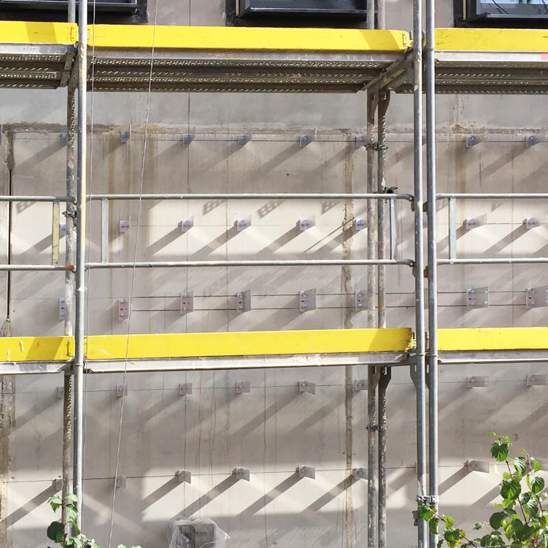 Eine Baustelle mit einem Baugerüst vor einer Fassade. An der Fassade sind mehrere Befestigungspunkte für Fassadendämmungen sichtbar. Das Gerüst ist sicher an der Fassade befestigt, um Arbeiten zu ermöglichen.