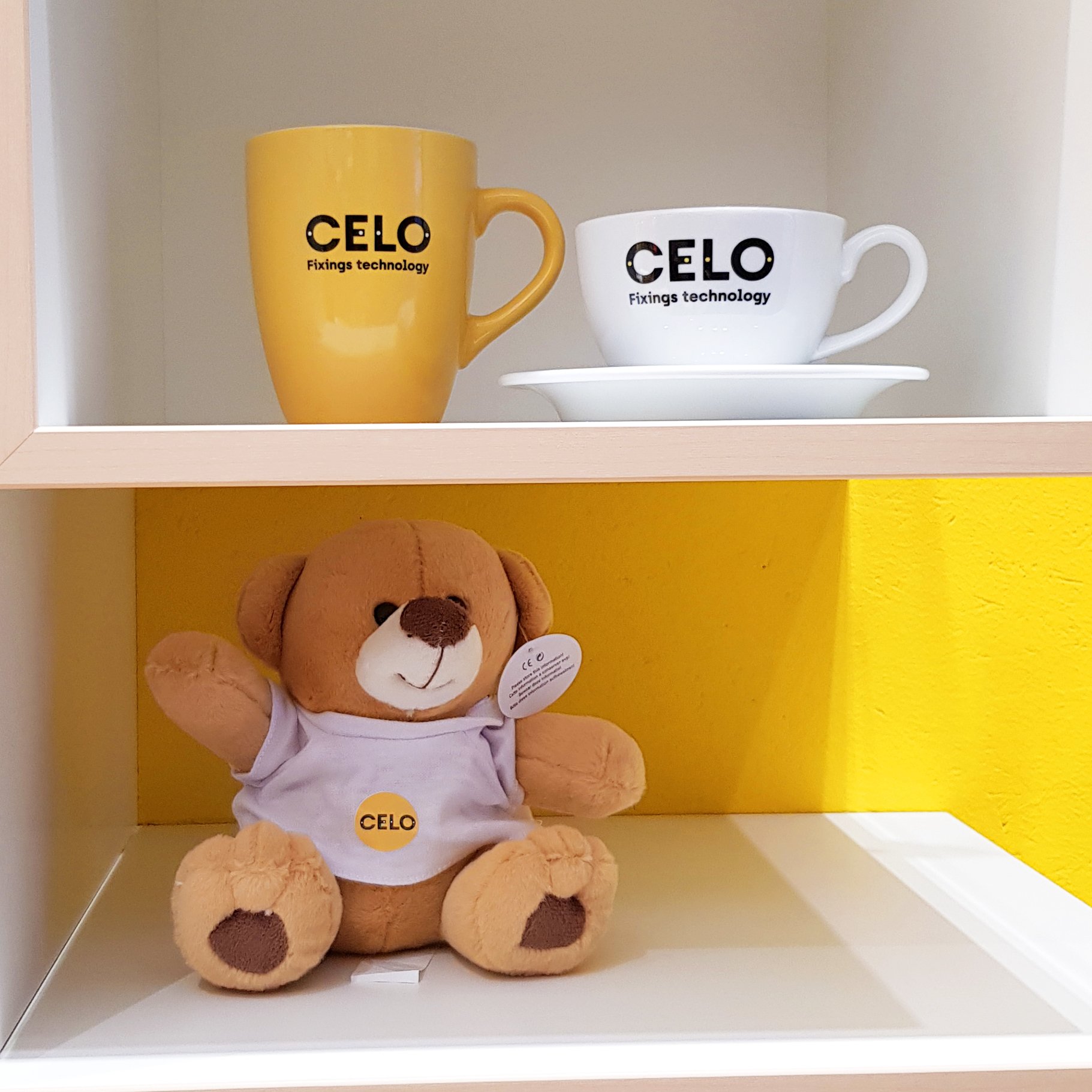 Teddybär und Tassen mit CELO-Logo in einem Regal